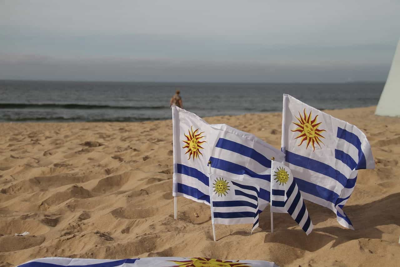 Cuatro pequeñas banderas de Uruguay clavadas en la arena de una playa, con mar de fondo.