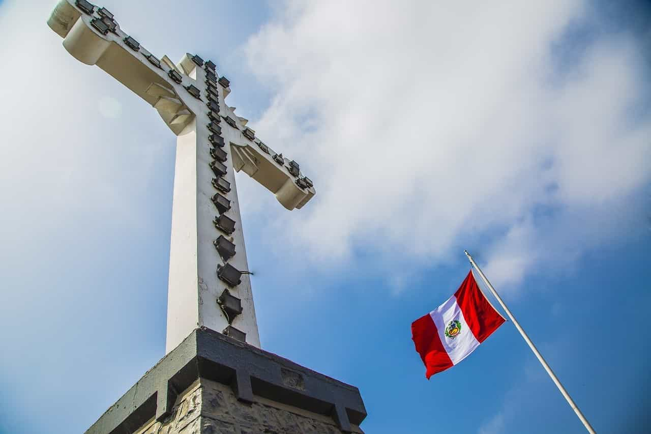 Bandera de Perú ondeando desde mástil junto a enorme monumento con forma de cruz católica.