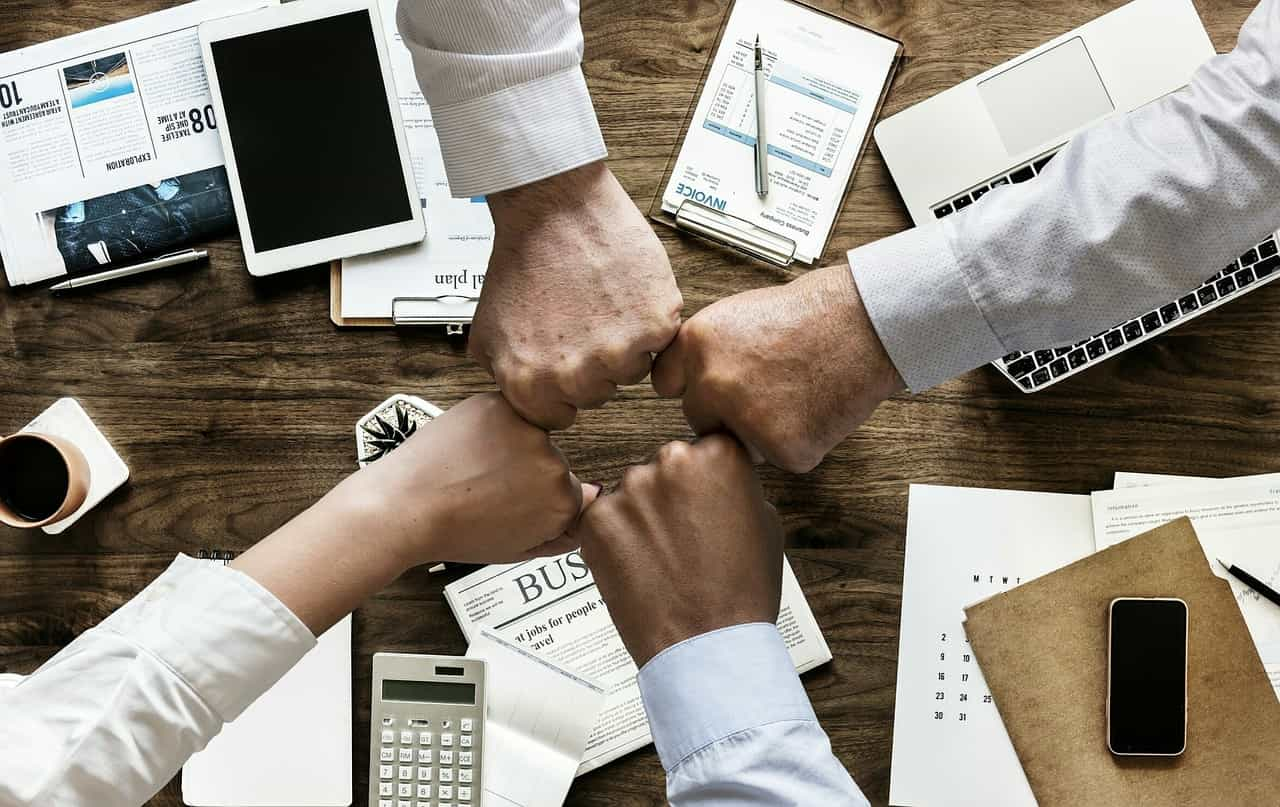 Puños de cuatro personas encontrándose en el centro de una mesa de trabajo sobre la que se despliegan distintos elementos de oficina.