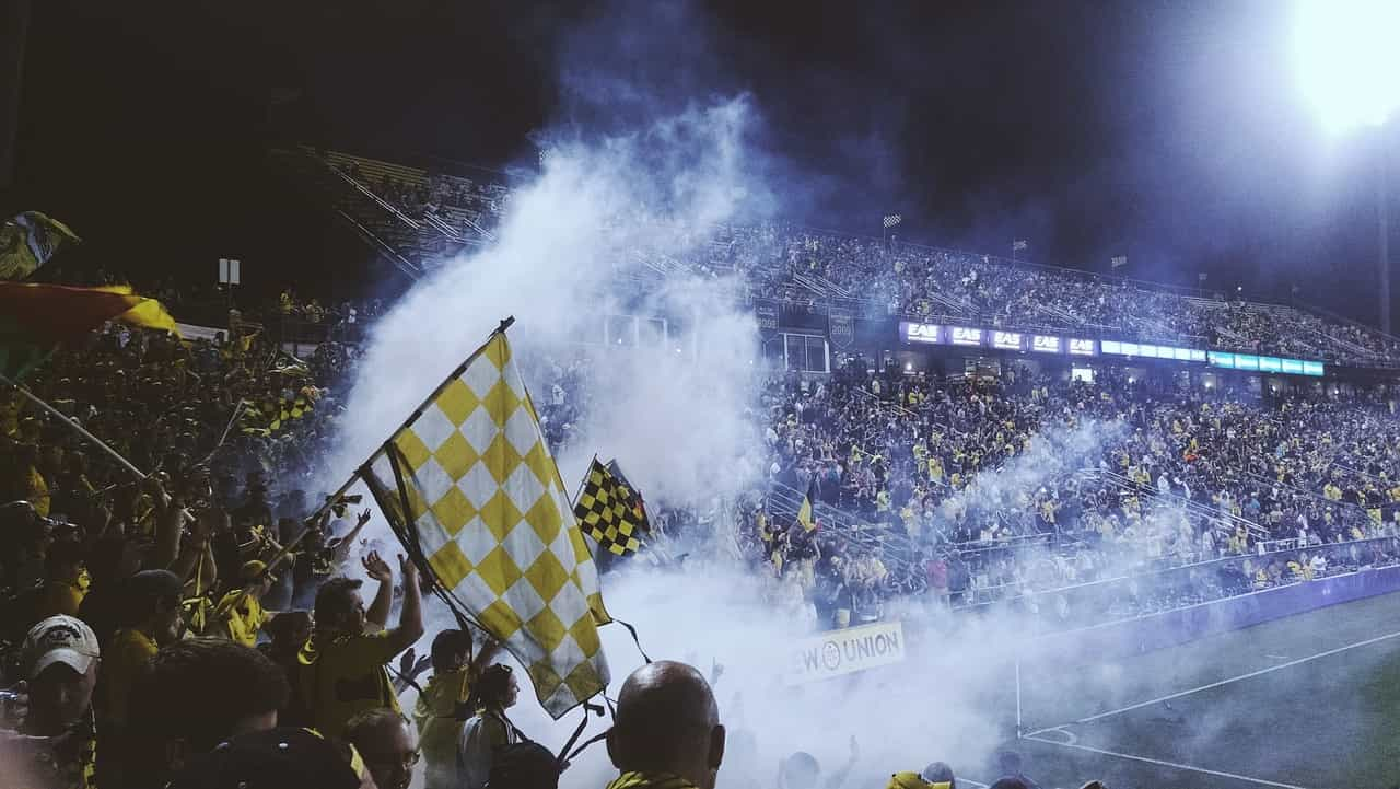 Estadio de fútbol colmado de hinchas que cantan, tocan bombos, encienden bengalas y agitan banderas.