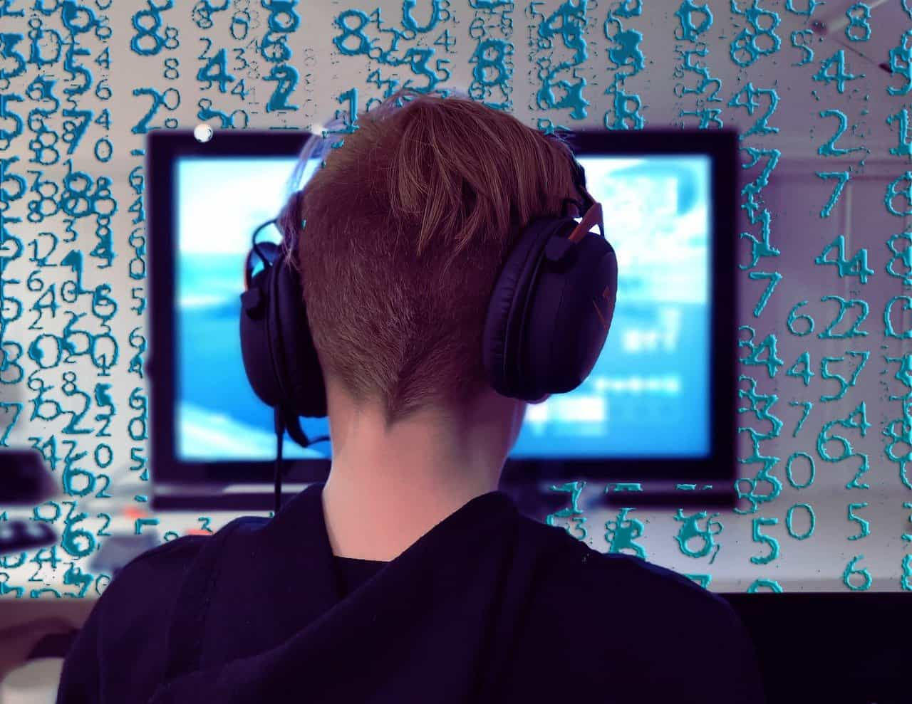 Joven de espaldas con auriculares puestos frente a una pantalla y una pared donde se despliega código informático.