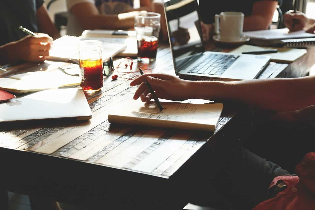 Personas sentadas a una mesa, tomando notas en cuadernos, con computadoras, vasos de refresco e insumo de oficina.