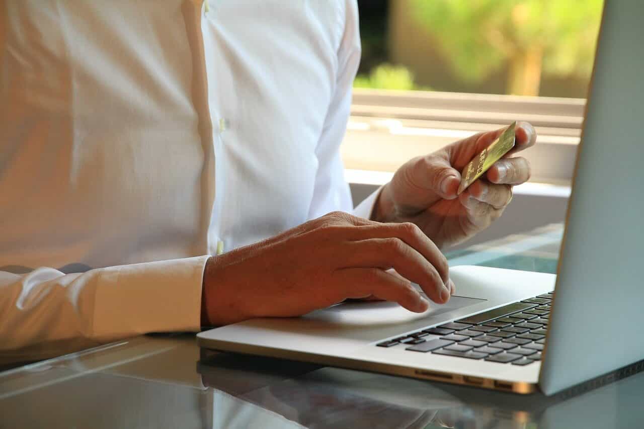 Persona vestida con camisa blanca frente a su ordenador portátil, con una tarjeta de crédito en la mano, en actitud de estar pagando online.