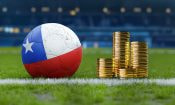 Balón de fútbol con los colores de la bandera de Chile junto a una pila de monedas en un campo de fútbol.